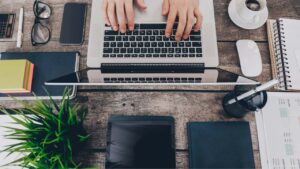 Top-down view of a wooden desk with a laptop, tablet, smartphone, glasses, plant, notebook, and coffee cup scattered around.